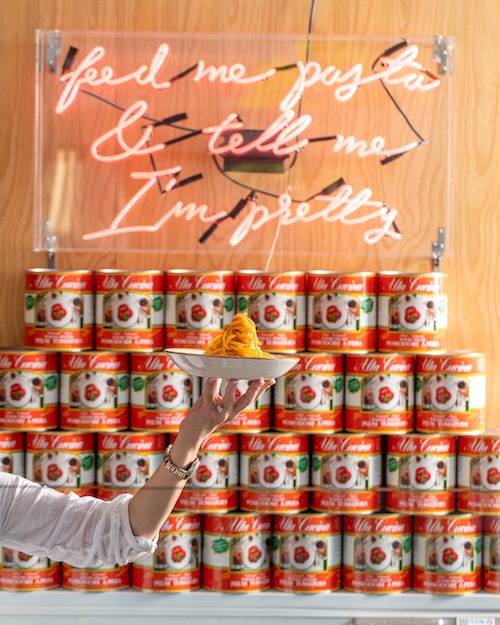Pasta dish in front of San Marzano tomato cans and a neon sign that says "& tell me I'm pretty"